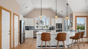 Een moderne keuken met witte kasten, roestvrijstalen apparatuur, een centraal eiland met barstoelen en uitzicht op een woongebied door grote ramen.