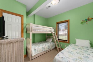 Une chambre avec un lit superposé, un téléviseur posé sur un meuble, une fenêtre avec des stores et un élément décoratif mural.