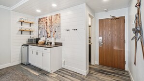 A modern kitchen with white cabinetry, a microwave, and a refrigerator. There is a wooden door and a ski rack on the wall.