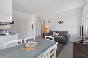 A modern kitchen with white cabinets, a dining table with chairs, a grey sofa, and a TV on a wooden stand.