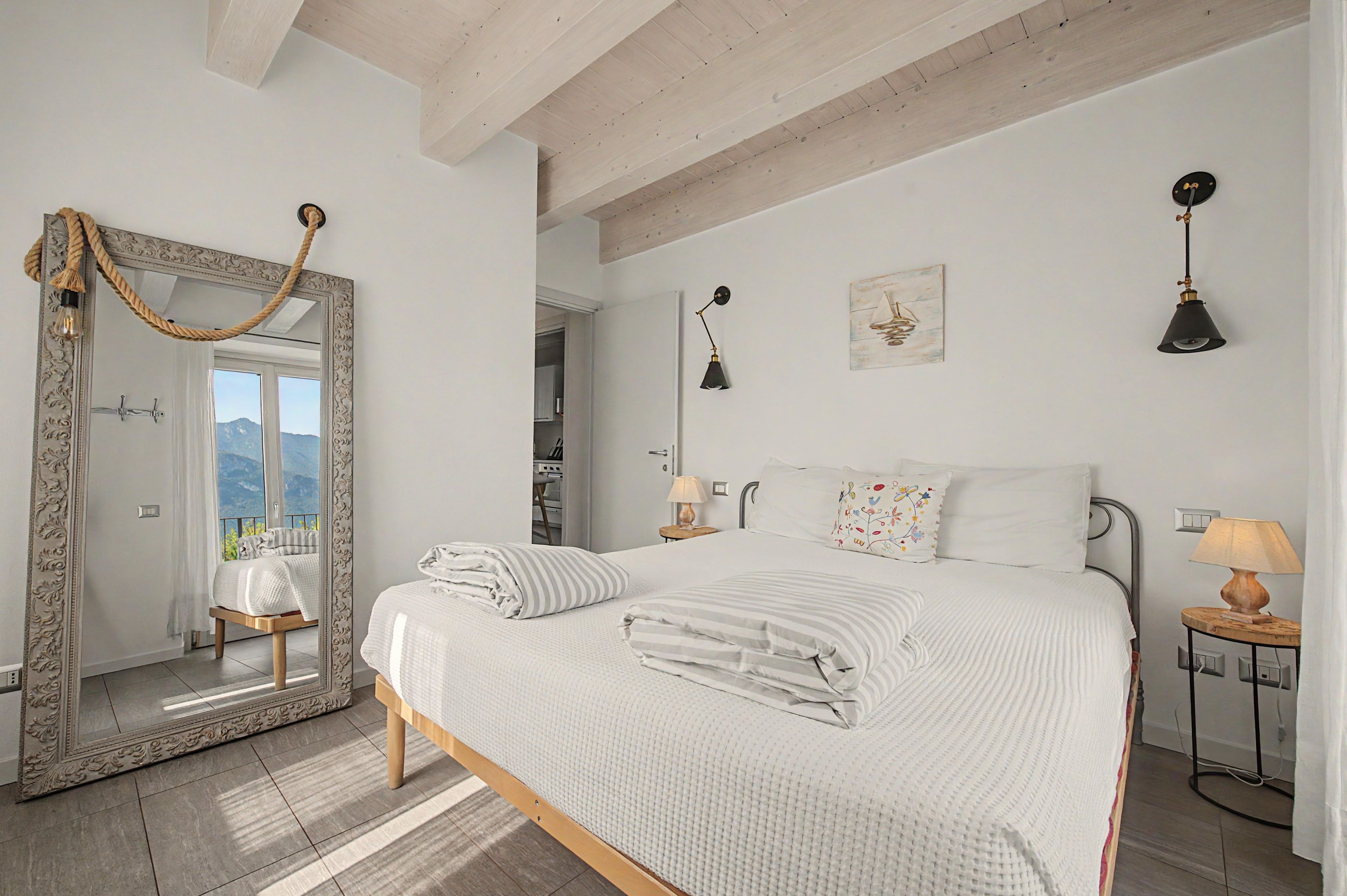 Un dormitorio con un espejo grande, una cama con ropa de cama blanca, una mesita de noche con lámpara y vistas a las montañas a través de la ventana.