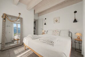 A bedroom with a large mirror, a bed with white bedding, a nightstand with a lamp, and a view of mountains through the window. - Apartment 'Aurora' with Lake View, Wi-Fi and Air Conditioning (Bellagio)