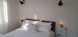Une chambre à coucher avec un lit, du linge de lit blanc, un coussin décoratif, des lampes de chevet et une étagère fixée au mur sur laquelle est posée une bougie.