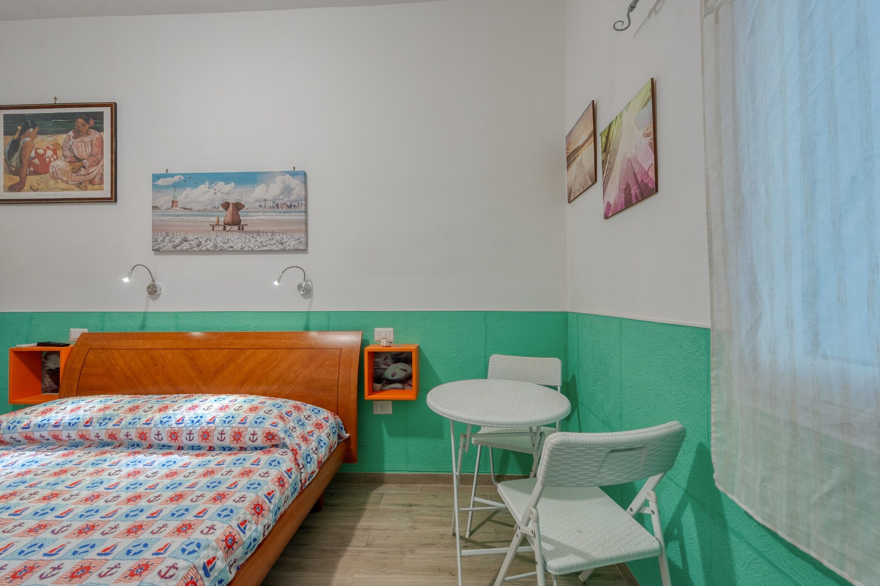 Ein Zimmer mit einem Bett, einem kleinen Tisch, einem Stuhl, einer Ablage mit einem Bild und einer Wand mit drei gerahmten Kunstwerken.