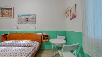 Ein Zimmer mit einem Bett, einem kleinen Tisch, einem Stuhl, einer Ablage mit einem Bild und einer Wand mit drei gerahmten Kunstwerken.