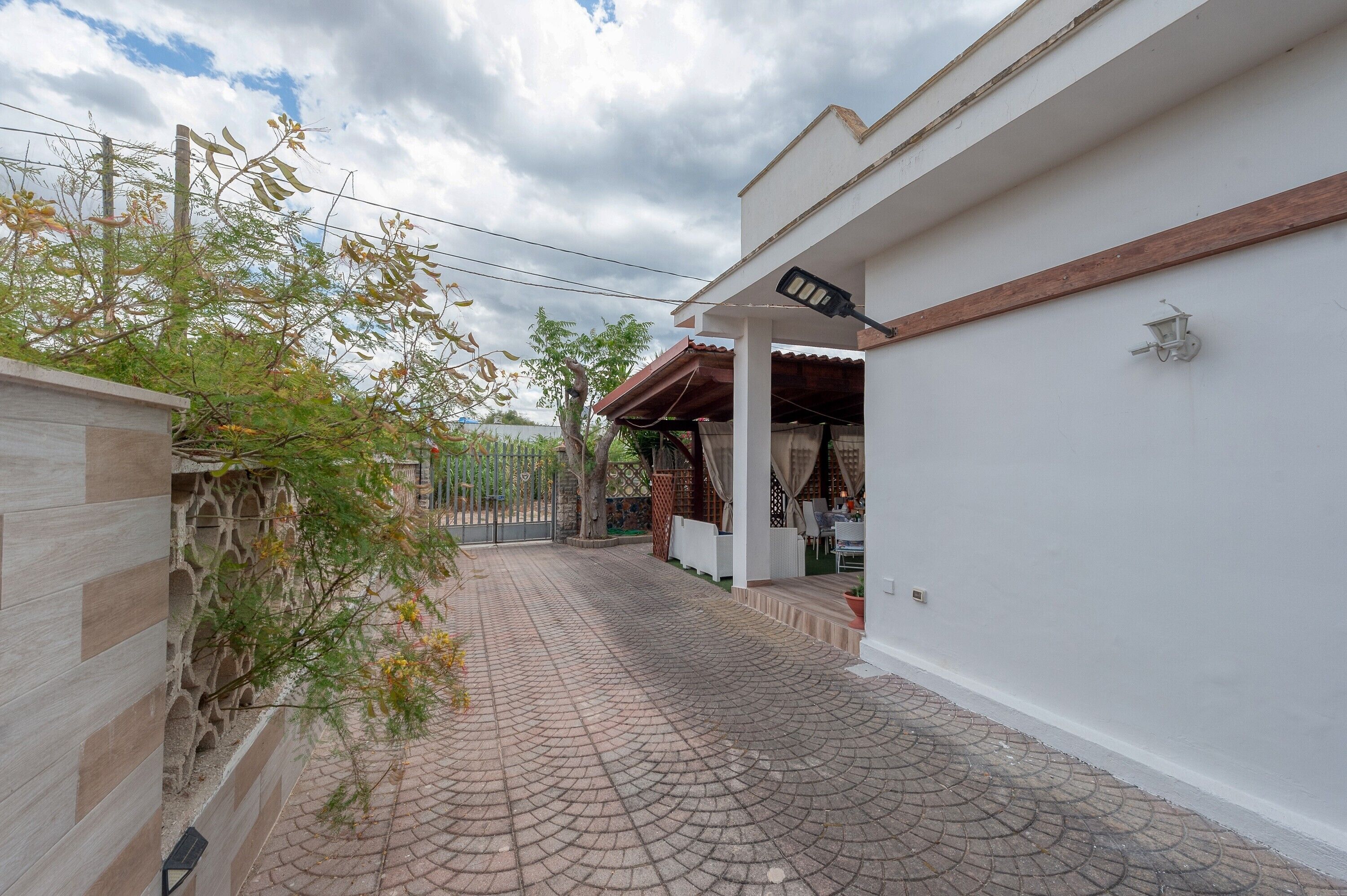 A paved pathway leading to a covered outdoor seating area with a white wall and a wooden beam.