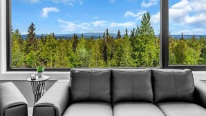 A modern living room with a grey sofa, a small round table with a plant, and large windows offering a view of a forest and mountains. - Mountain Lake Sanctuary in Alaska Up to 22 Guests (Anchorage)