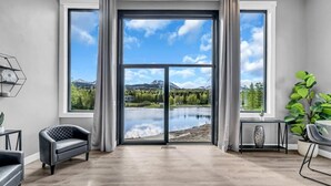 A room with large windows, a view of a lake and mountains, a black leather chair, a small table with a vase, and a potted plant. - Mountain Lake Sanctuary in Alaska Up to 22 Guests (Anchorage)