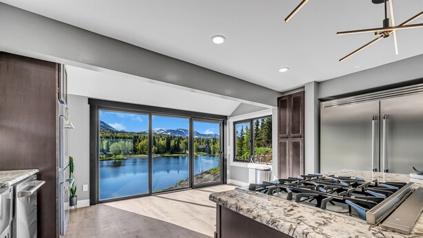 A modern kitchen with a large island, a stainless steel refrigerator, and a view of a lake and mountains through large sliding glass doors. - Mountain Lake Sanctuary in Alaska Up to 22 Guests (Anchorage)