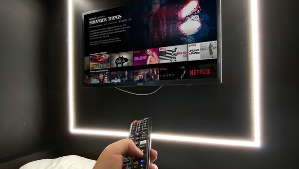 A person holding a remote control in front of a wall-mounted TV displaying a Netflix interface. - Habitación  Tipo Cápsula (Bogota)