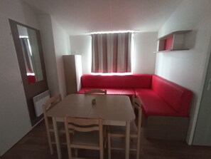 Une petite pièce avec un canapé rouge, une table en bois entourée de chaises, un miroir et une fenêtre avec des rideaux.