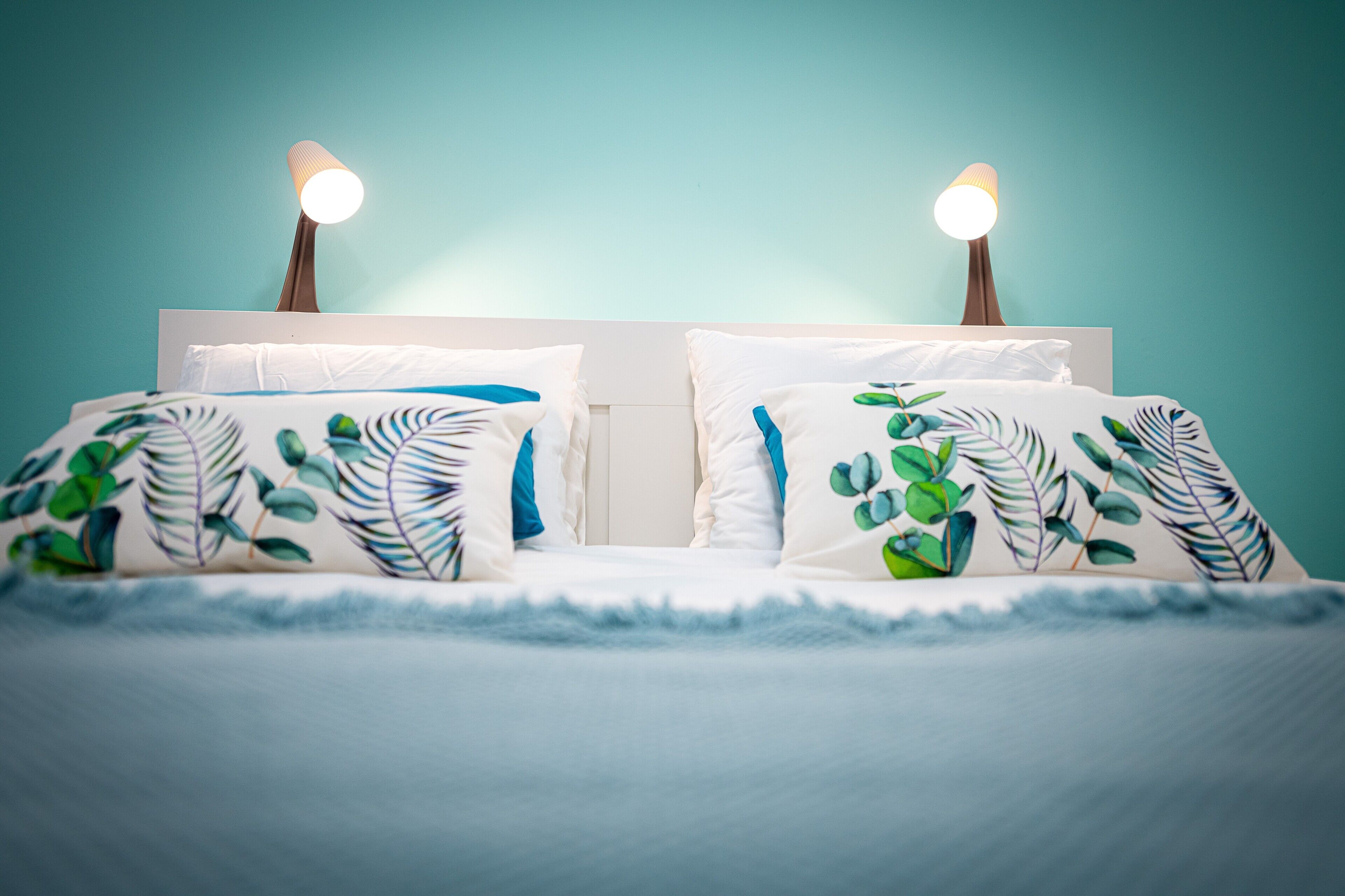A bed with white bedding and leaf-patterned pillows, two bedside lamps, and a teal wall.
