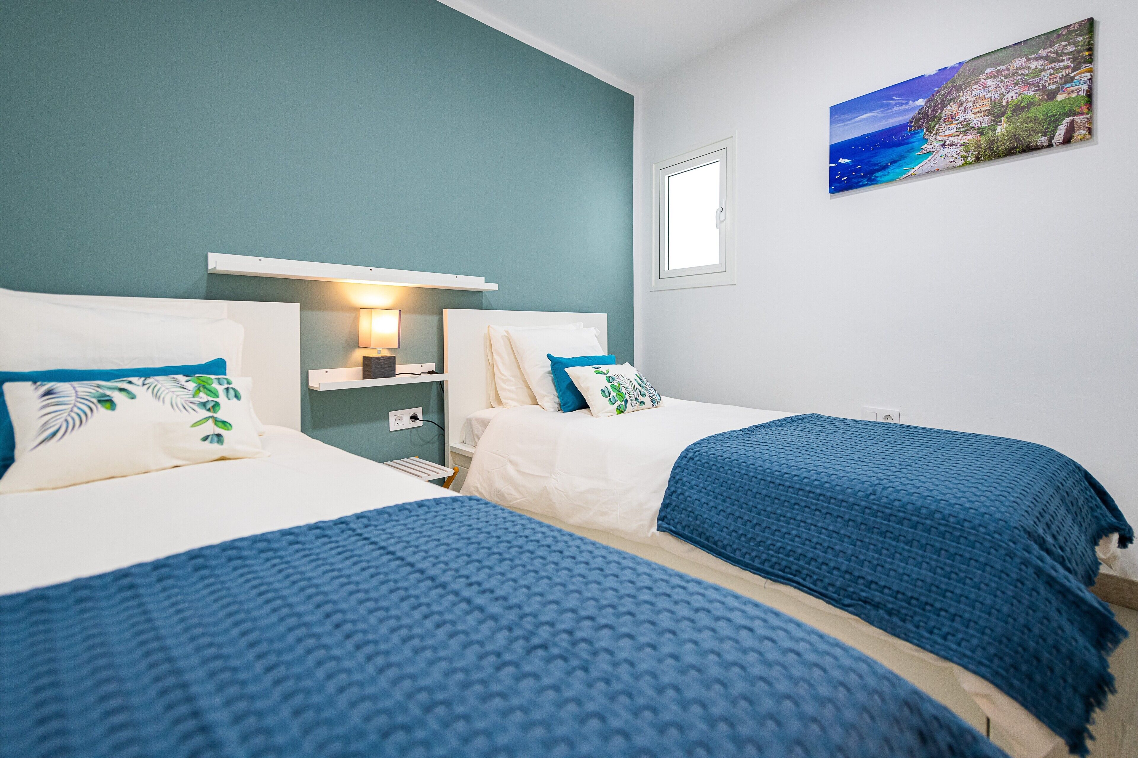 A bedroom with two beds, a nightstand, a lamp, and a painting of a coastal scene.