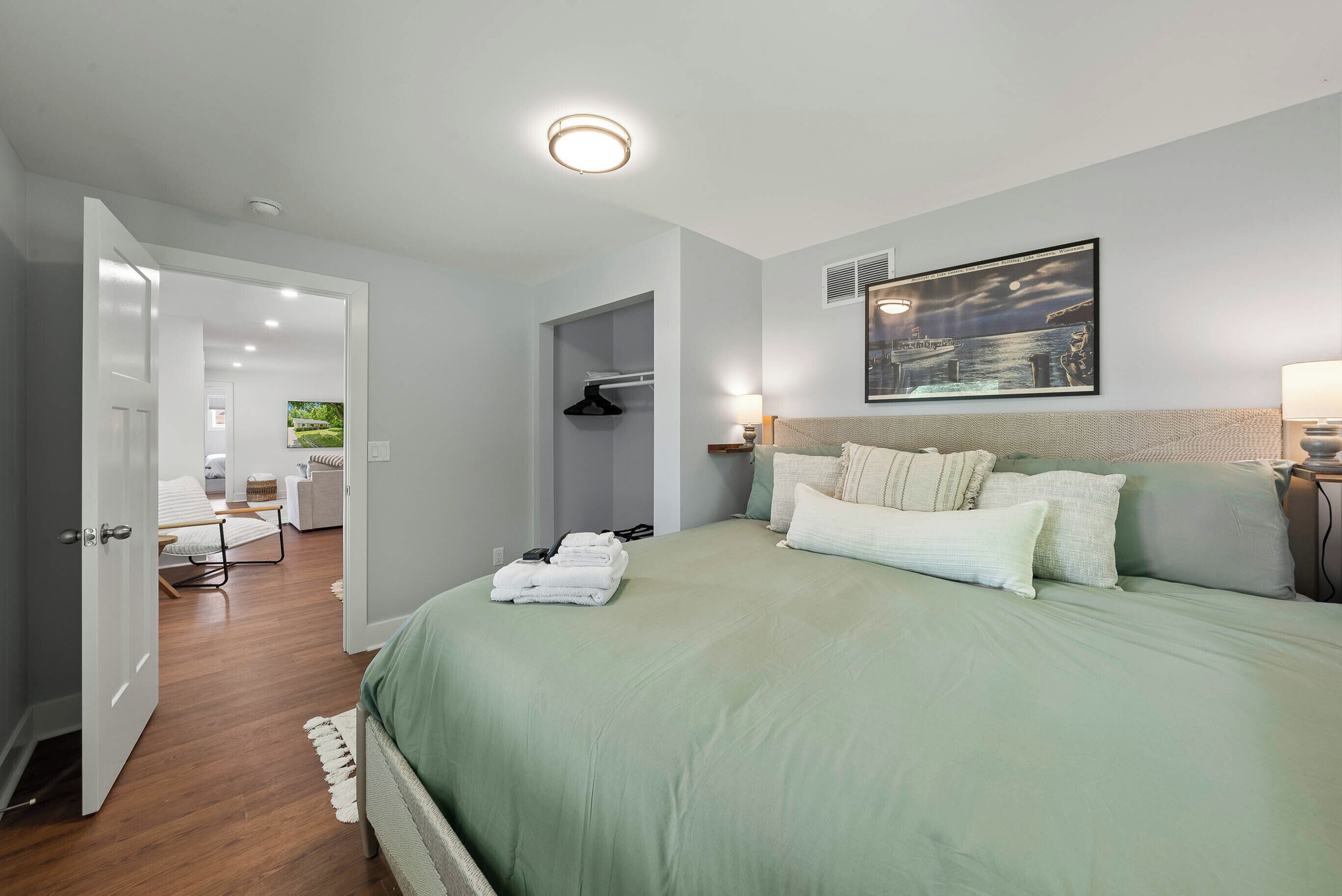 A bedroom with a large bed, a nightstand, a wall-mounted painting, and a door leading to a bathroom.