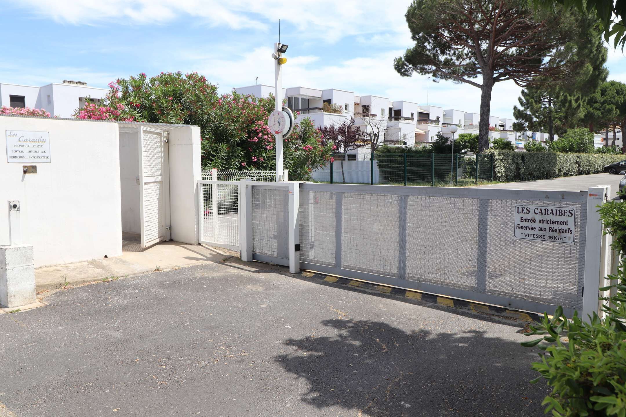 Une entrée fermée par un portail donne accès à un quartier résidentiel. Un panneau indique « LES CARAIRES », et un indicateur de distance signale « 15 km ».