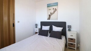 A bedroom with a bed, two bedside tables, a nightstand, a wooden wardrobe, and a beach-themed wall art.