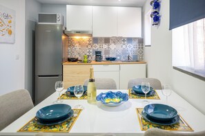 A compact kitchen with a refrigerator, microwave, and coffee maker. A dining table with blue plates and a bottle of white wine is set for two.