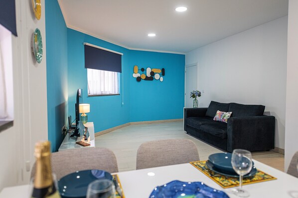 A modern living room with a blue accent wall, a black sofa, a dining table with chairs, and a decorative wall piece.