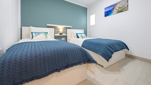 Une chambre avec deux lits, une table de chevet, une lampe fixée au mur et un tableau représentant un paysage marin.