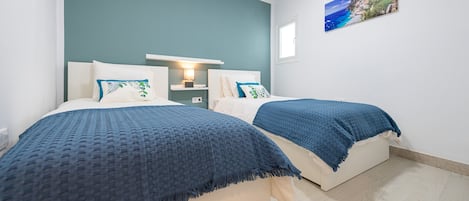 A bedroom with two beds, a nightstand, a wall-mounted lamp, and a coastal view painting.