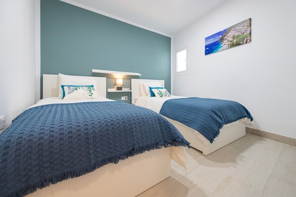 A bedroom with two beds, a nightstand, a wall-mounted lamp, and a coastal view painting.
