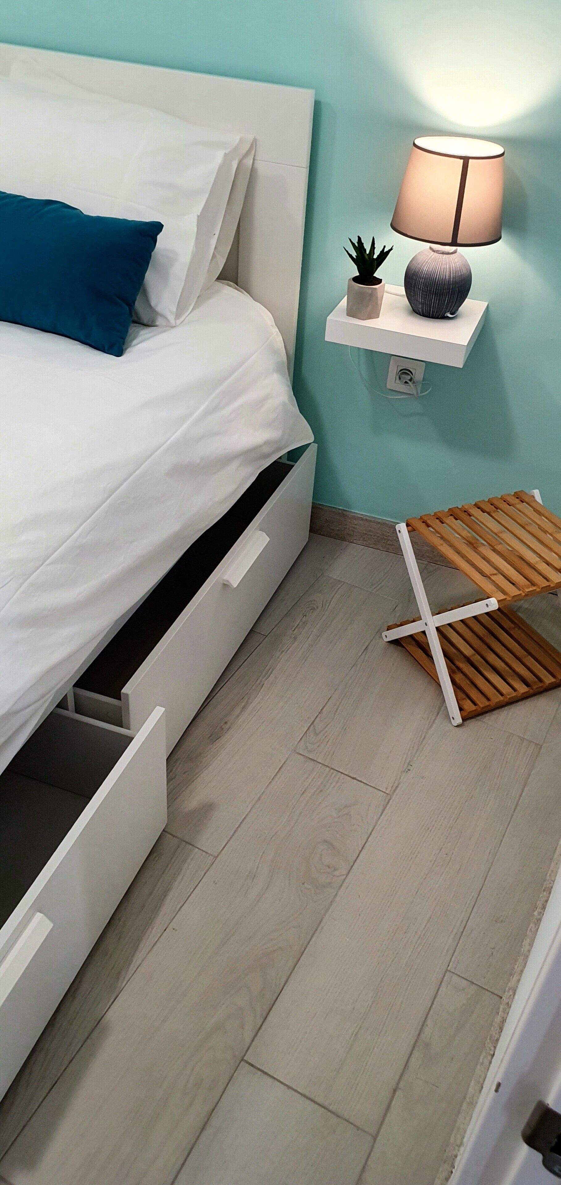 Un dormitorio con una cama, una mesita de noche con una lámpara, una planta pequeña y un taburete de madera.