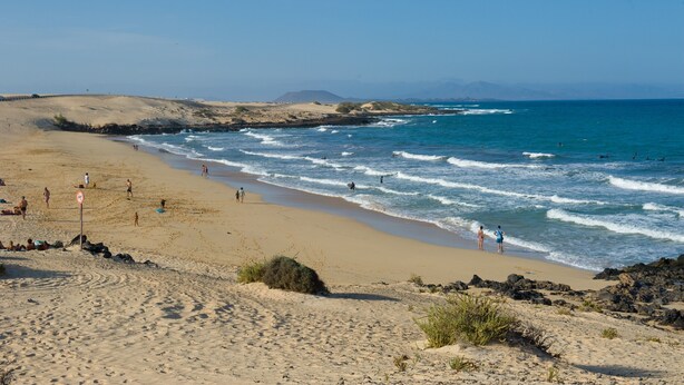 A sandy beach with waves, a clear sky, and distant mountains.