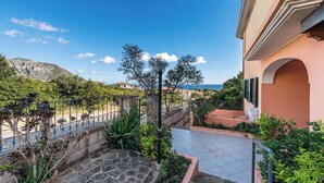 A balcony with a stone floor, potted plants, and a view of the sea and mountains. - Holiday home with veranda in the inner courtyard (Cala Gonone)