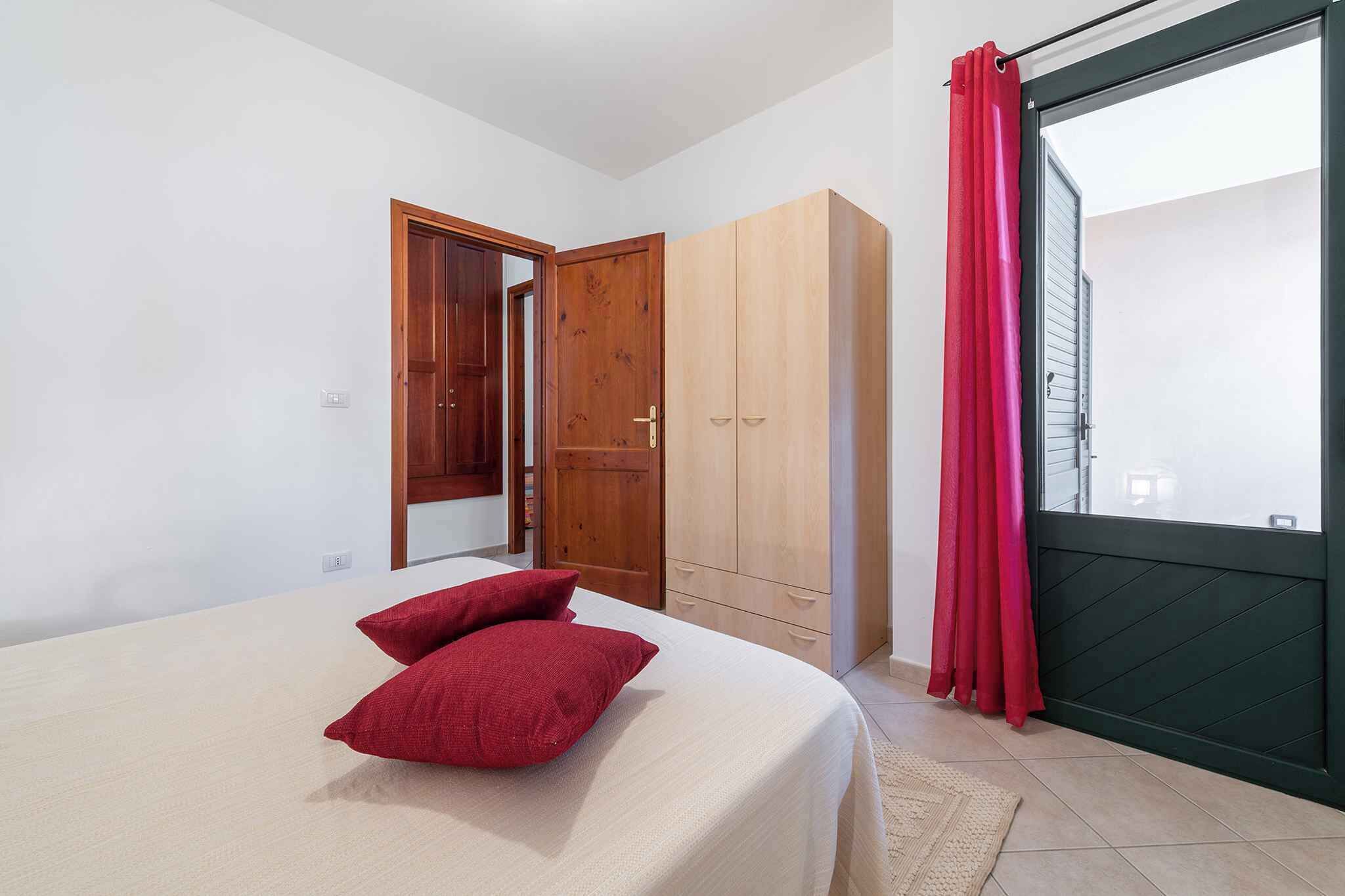 Une chambre à coucher avec un lit, des oreillers rouges, une armoire et une porte coulissante donnant sur un balcon.