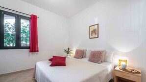 A bedroom with a bed, a nightstand, a window with a red curtain, a wall-mounted picture, and a bedside lamp. - Holiday home with veranda in the inner courtyard (Cala Gonone)