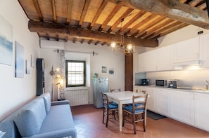A compact kitchen with white cabinets, a dining table with chairs, a built-in oven, and a window with curtains.