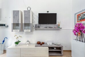 Un soggiorno moderno con una TV a schermo piatto montata su una console grigia, un armadietto bianco con ante in vetro, un tavolino con oggetti decorativi e una pianta di orchidea in vaso.