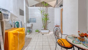 Un pequeño patio cubierto con suelo de baldosas, una planta en maceta, una mesa techada y una silla.