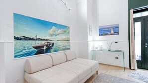 Una habitación con un cuadro grande de paisaje marino, un sofá, una mesa de vidrio y una puerta con cortinas.