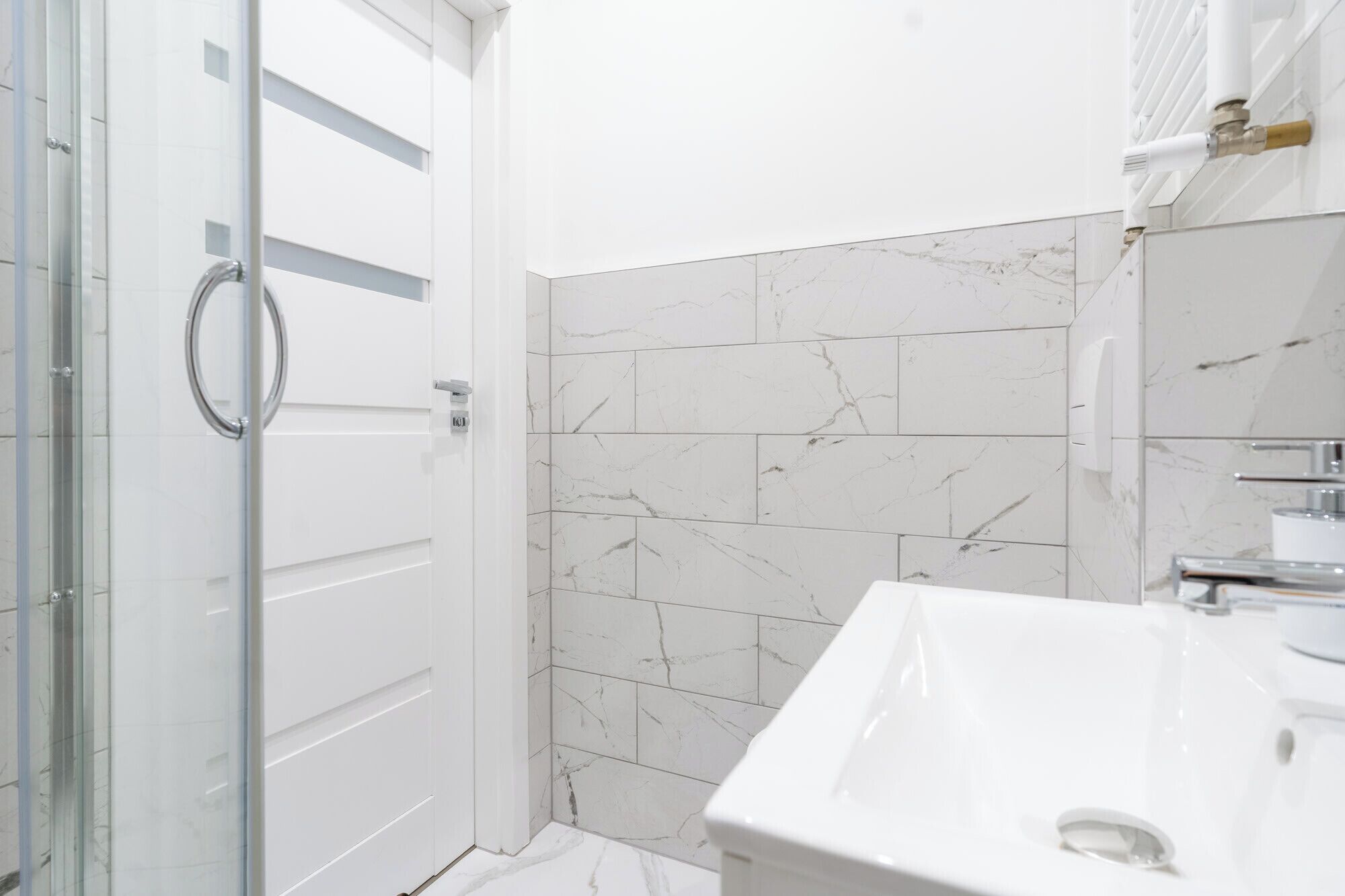 Un baño moderno con mampara de vidrio, azulejos de mármol blanco y lavamanos blanco.