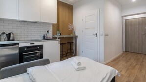 A modern hotel room with a kitchenette, a bed with a rolled towel, and a wooden door.