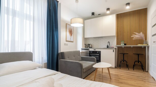 A modern hotel room with a bed, a grey sofa, a small white table, and a kitchenette in the background.