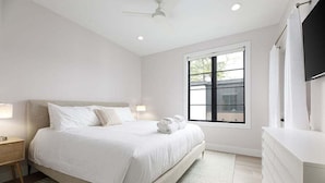Un dormitorio moderno con una cama grande, mesitas de noche, ventilador de techo, ventana y televisor montado en la pared.