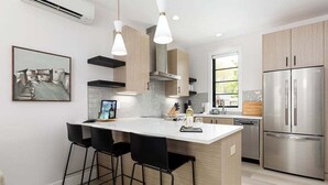 Une cuisine moderne dotée d’un îlot central, de tabourets de bar noirs, d’appareils électroménagers en acier inoxydable et d’une grande fenêtre qui laisse entrer la lumière naturelle.