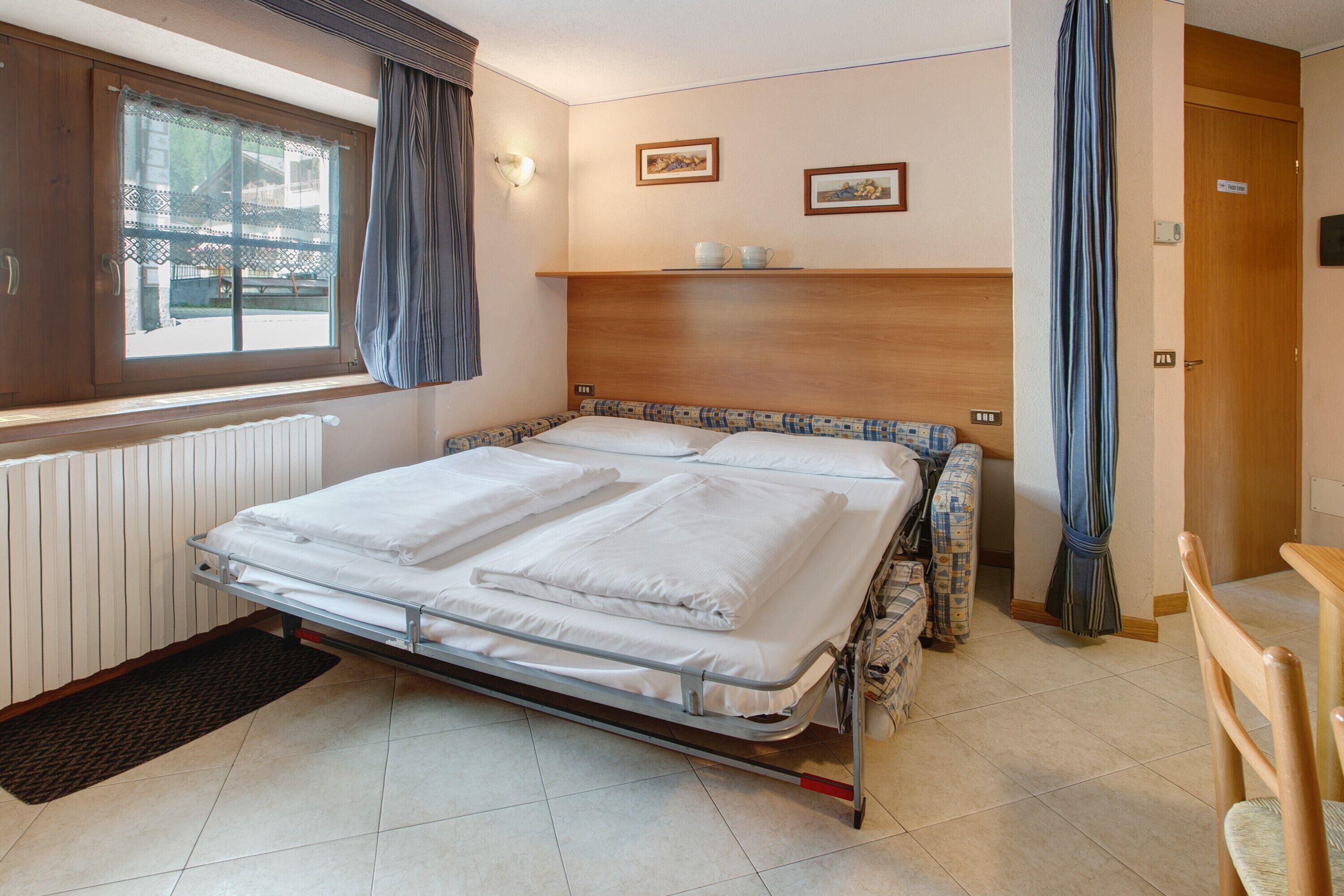 A hotel room with a bed, a window with curtains, a radiator, a wooden headboard, and a small shelf with cups.