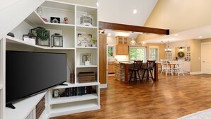 A spacious living area with a large flat-screen TV, wooden flooring, a dining area with a kitchen in the background, and a staircase with white shelving. - Maple City Farm | Pet Friendly (Maple City)