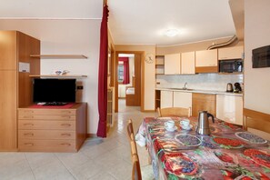 Una habitación pequeña con televisión, armarios de madera, una mesa de comedor con mantel estampado de frutas y una zona de cocina con microondas.