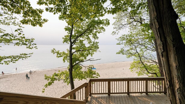 Une terrasse en bois surplombe une plage où l’on voit des gens se promener et un bateau sur l’eau.