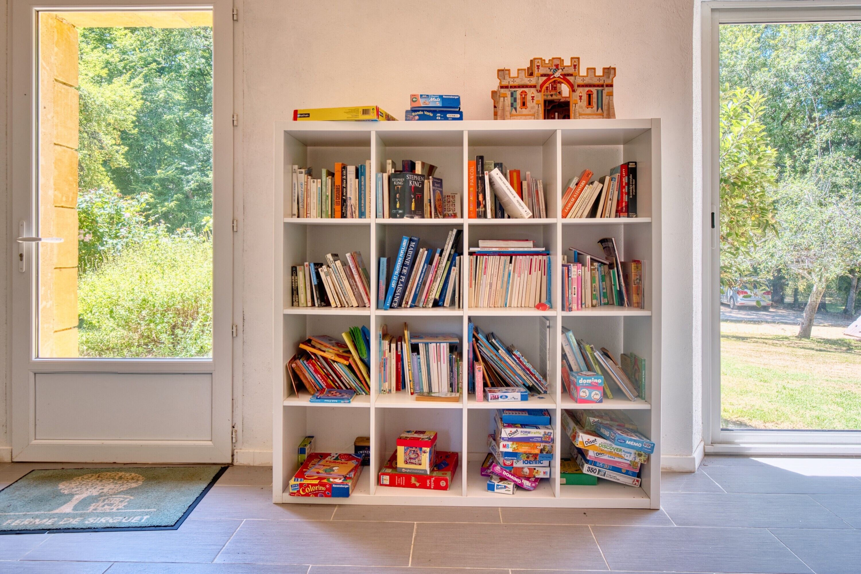 Une bibliothèque remplie de livres et de jouets, un petit modèle de château et un paillasson orné d’un motif d’arbre.