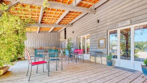 Una terraza de madera con mobiliario exterior, un espacio cubierto con techo de madera y la entrada de un edificio con un letrero.