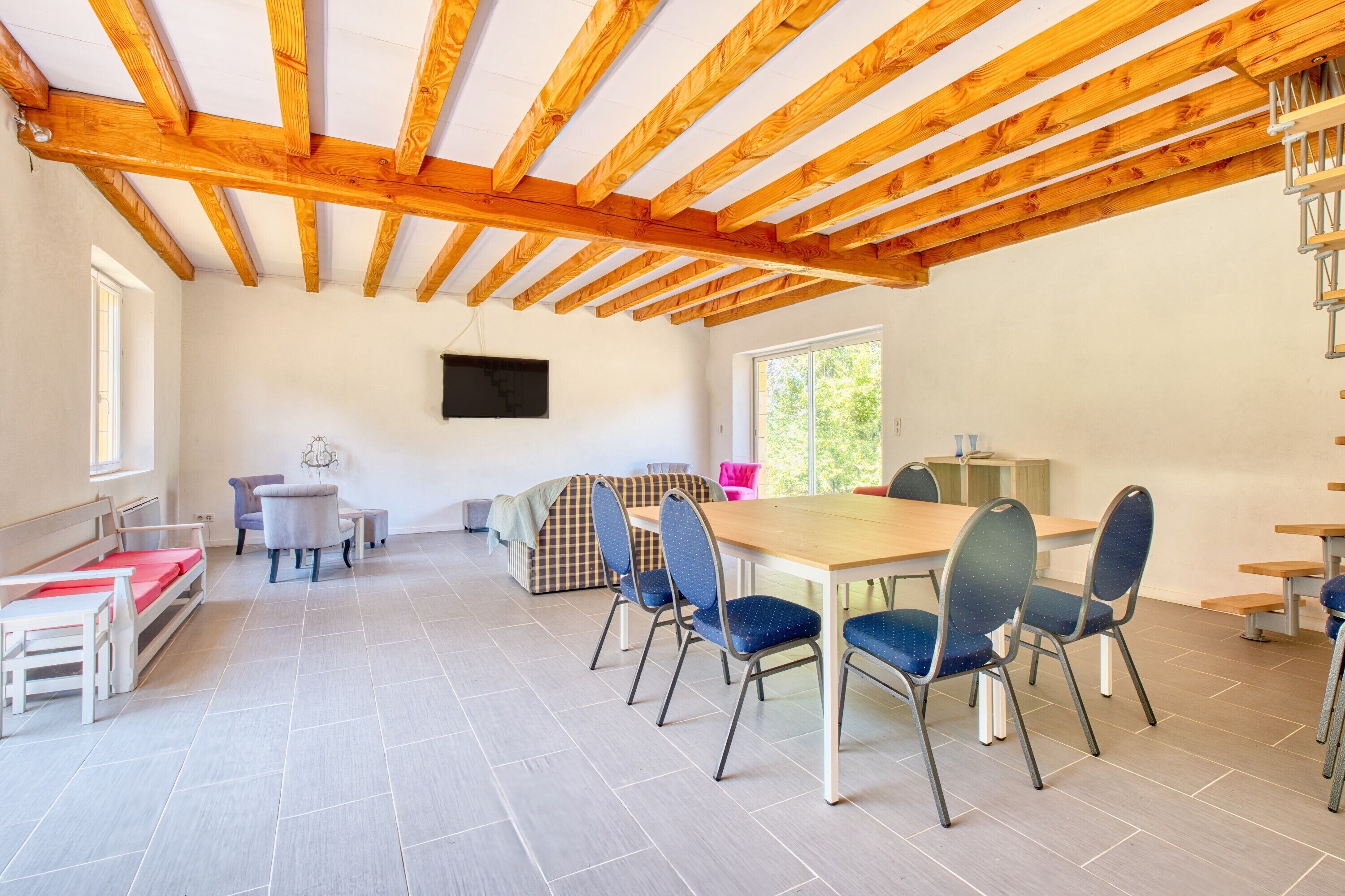 A spacious room with a wooden ceiling, a dining table with chairs, a sofa, and a television mounted on the wall.