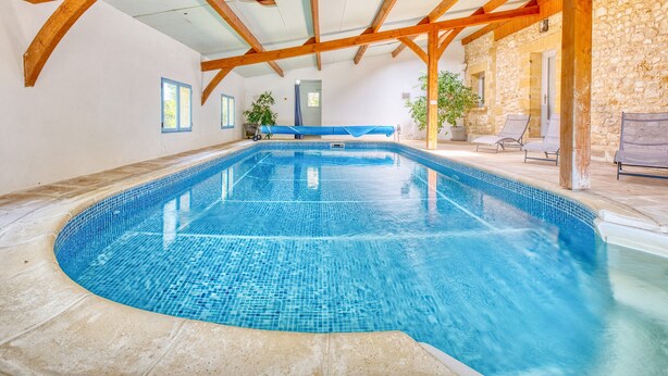 Una piscina interior con suelo de baldosas, vigas de madera, paredes de piedra y cielo azul visible a través del techo.