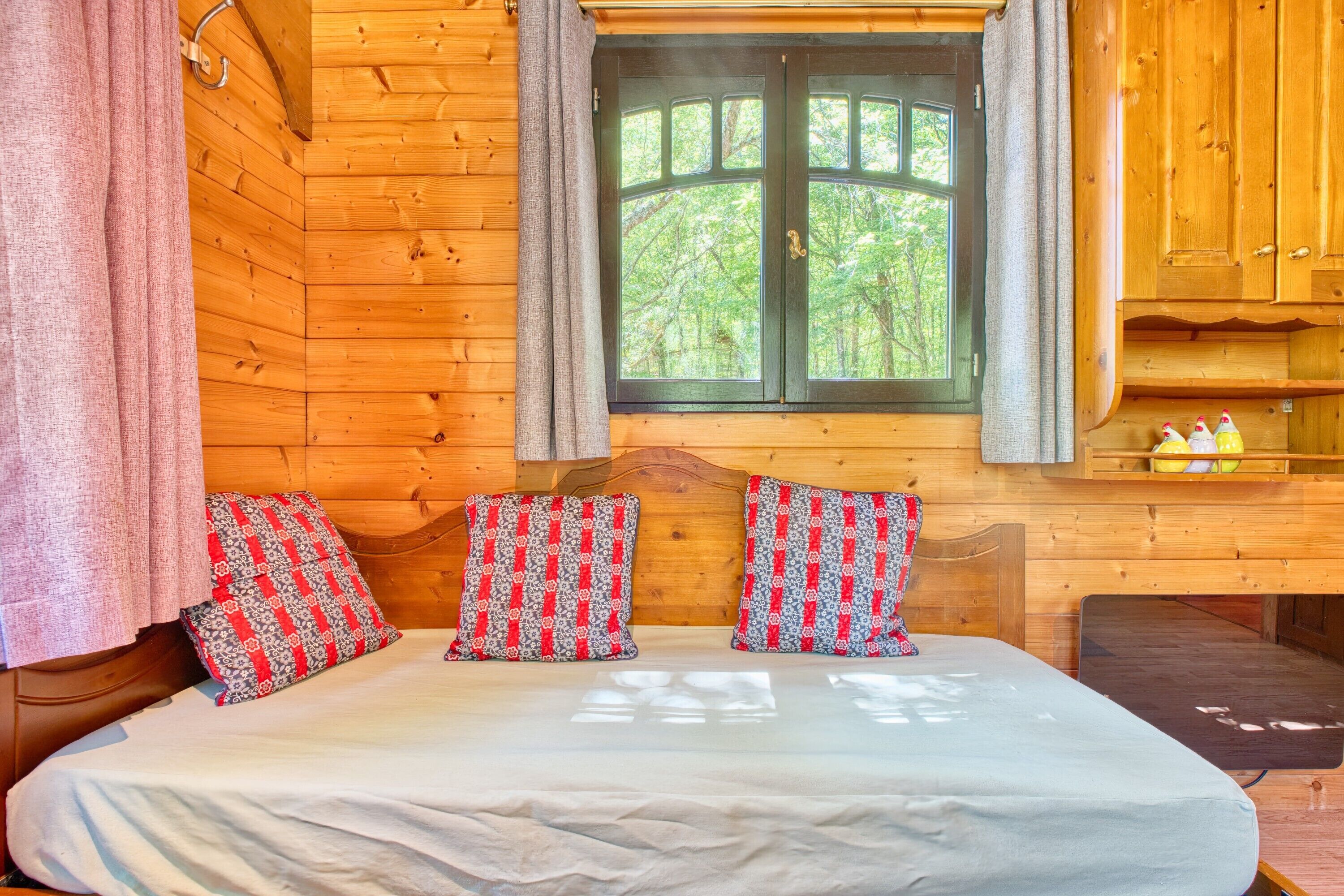 Une chambre rustique en bois, avec un lit, deux coussins à motifs, une fenêtre avec des rideaux et une armoire en bois contenant des bouteilles.