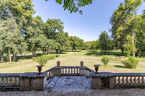 A courtyard with a stone balustrade, potted plants, and a grassy lawn surrounded by trees.