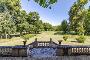A courtyard with a stone balustrade, potted plants, and a grassy lawn surrounded by trees.